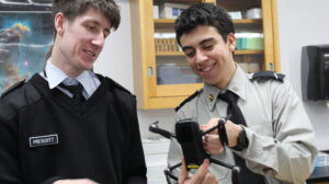 Two students in uniform smile while examining and adjusting a small drone together in a classroom setting.