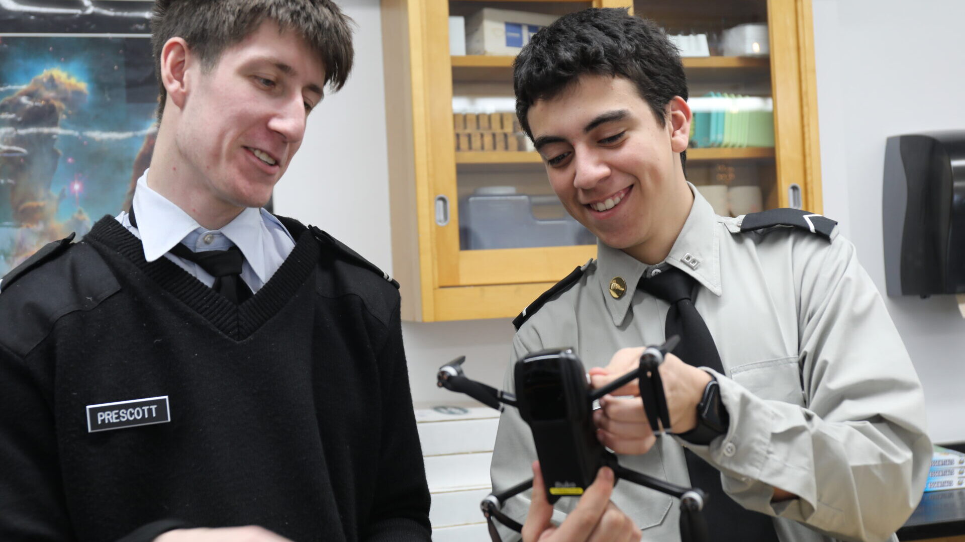 Two students in uniform smile while examining and adjusting a small drone together in a classroom setting.