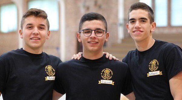 Three student cadets stand together indoors, smiling with arms around each other while wearing matching black academy T-shirts.