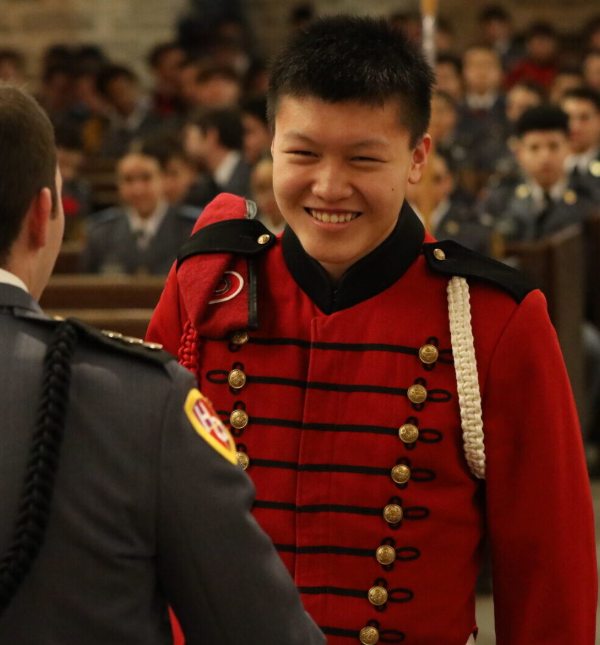 Cadets in dress uniforms exchange a handshake inside a chapel, one smiling in red while rows of seated cadets watch.