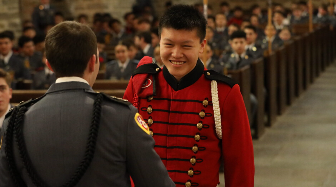 Cadets in dress uniforms exchange a handshake inside a chapel, one smiling in red while rows of seated cadets watch.