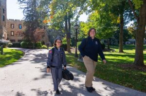 Two students walk along a tree-lined campus path, one in a cadet uniform and one in casual attire, smiling on a sunny day.