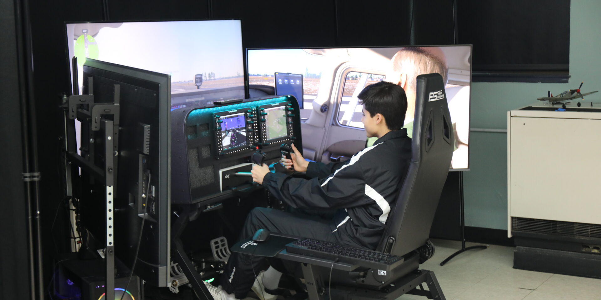 Student sits in a flight simulator cockpit using controls and screens to practice piloting an aircraft in a classroom lab.